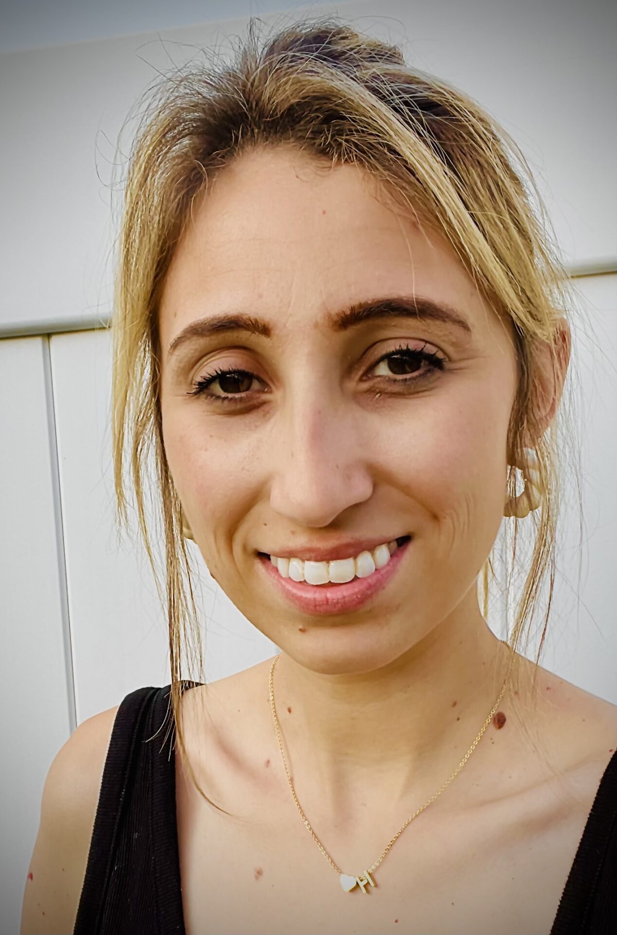 A woman with blonde hair tied back is smiling at the camera. She is wearing a black top, a delicate gold necklace with two small pendants, and small hoop earrings. The background shows a white fence.