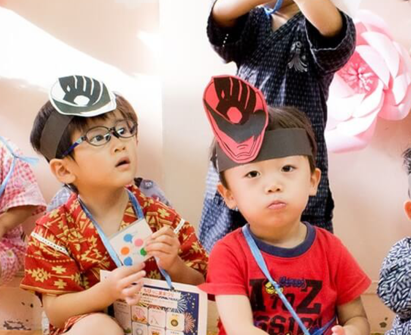 Two young boys are sitting and wearing black headbands with paper animal faces. The boy on the left wears glasses and a patterned shirt, and holds an envelope. The boy on the right is in a red shirt with a blue lanyard. Other children and decorations are in the background.